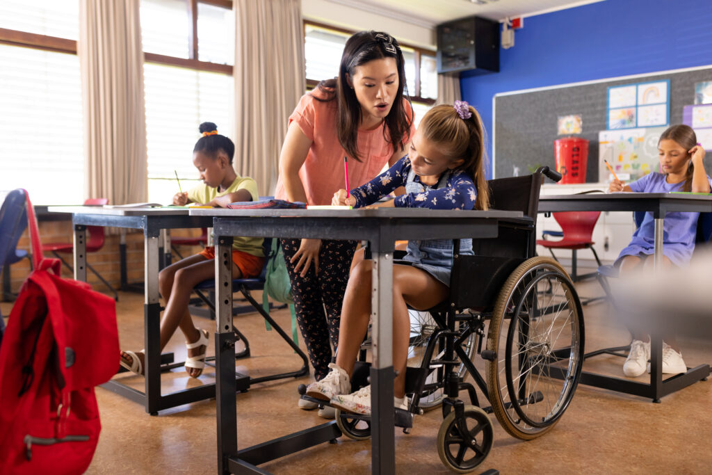 Teacher girl in a wheelchair at school, after class. They are in a colorful classroom, with other children focused on their activities.