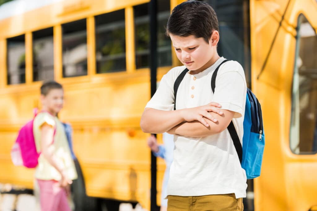 depressed little schoolboy with crossed arms standing in front of school bus
