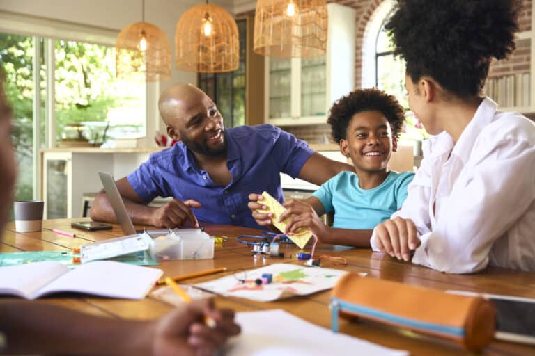 Family Around Table At Home Using Laptop With Parents Helping Children With Science Homework
