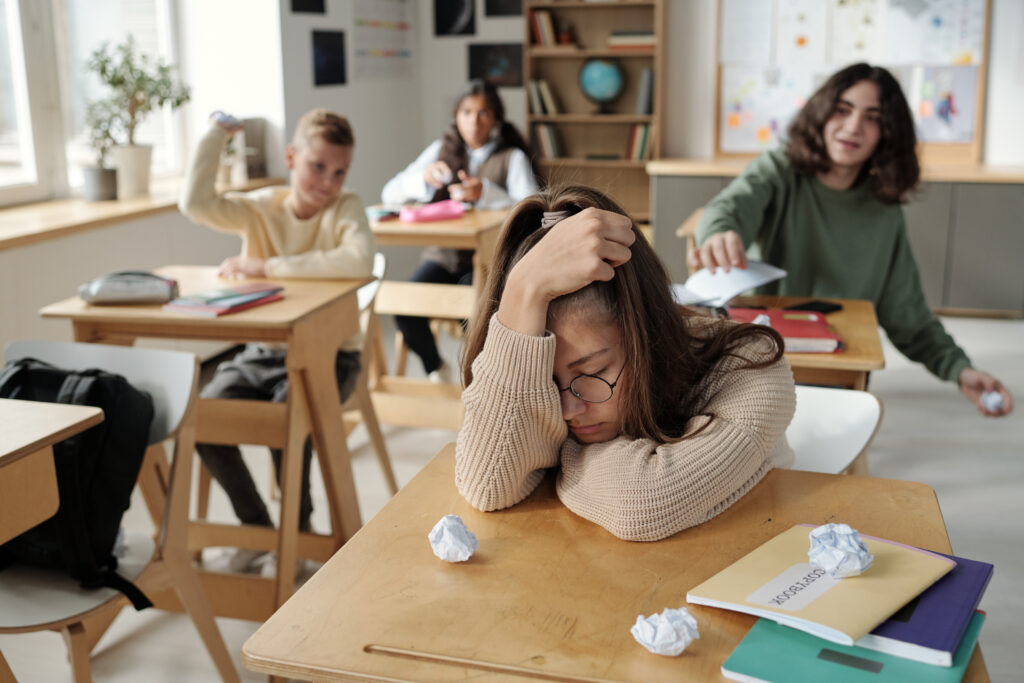 Childhood trauma concept with offended schoolgirl sitting by her desk while group of cruel classmates throwing crumpled papers at her