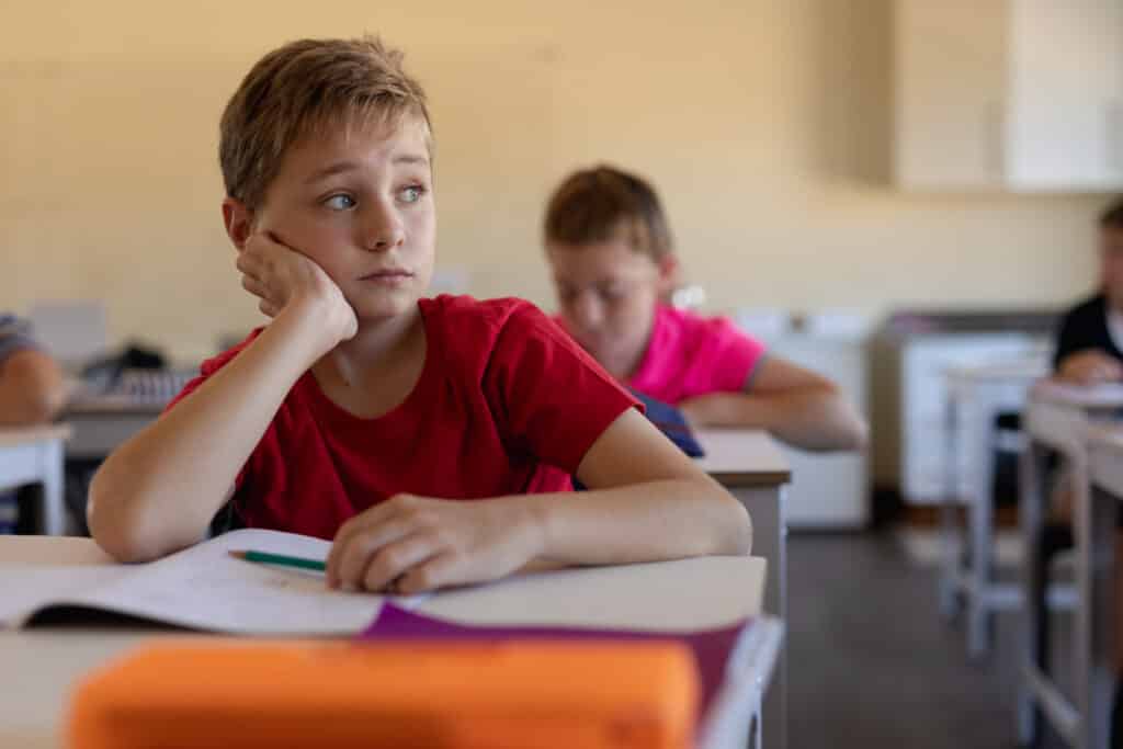 Front view close up of a Caucasian schoolboy with short hair wearing a red t shirt sitting at a desk in an elementary school classroom leaning on his hand and looking away, an open book on the desk in front of him, and classmates sititng and working in the background