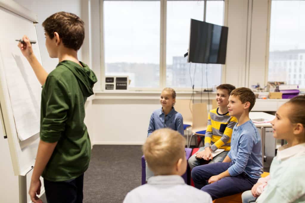 education, elementary school, learning and people concept - happy student boy with marker writing on flip board