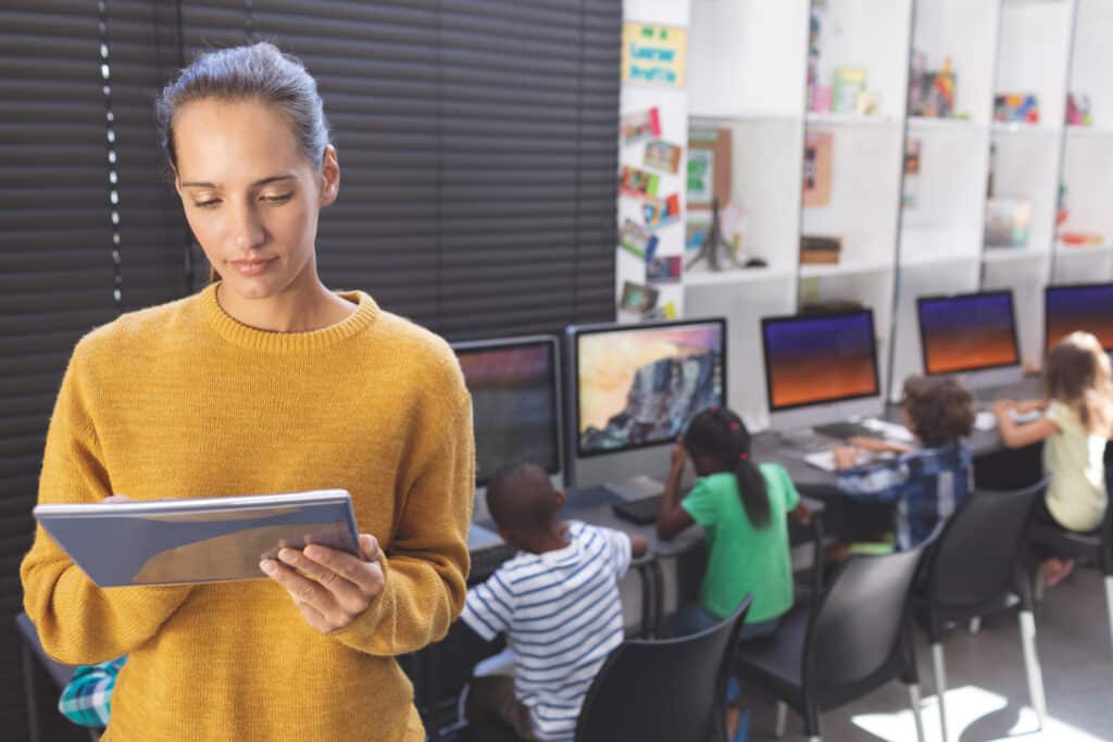 Teacher using digital tablet while kids working on computer behind her in computer room
