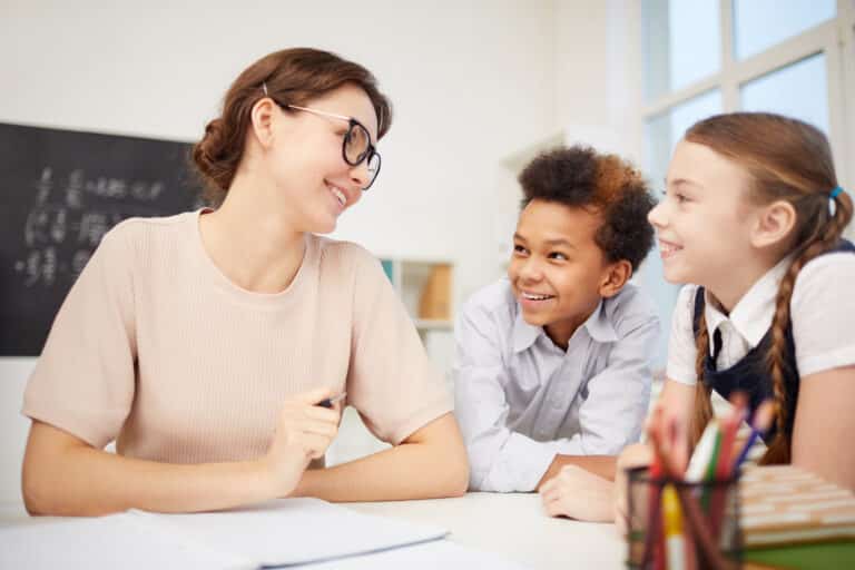 Young teacher in eyeglasses sitting at her workplace and smiling to children who talking to her social emotional learning concept