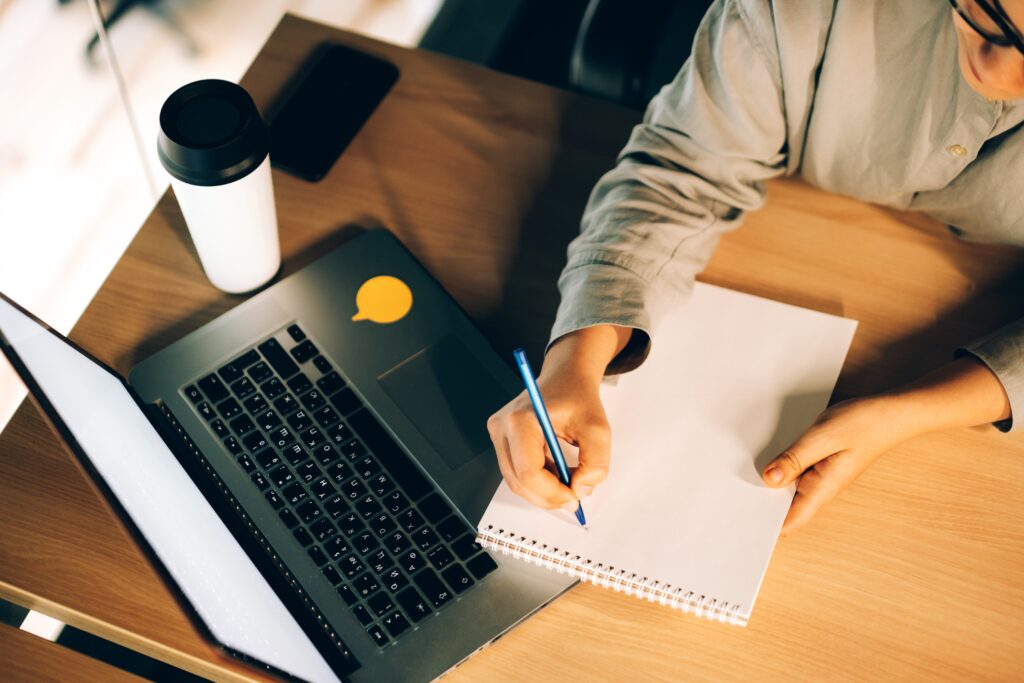 Young woman studying online teaching courses with a laptop, writing down notes in notebook and drinking coffee from reusable glass.